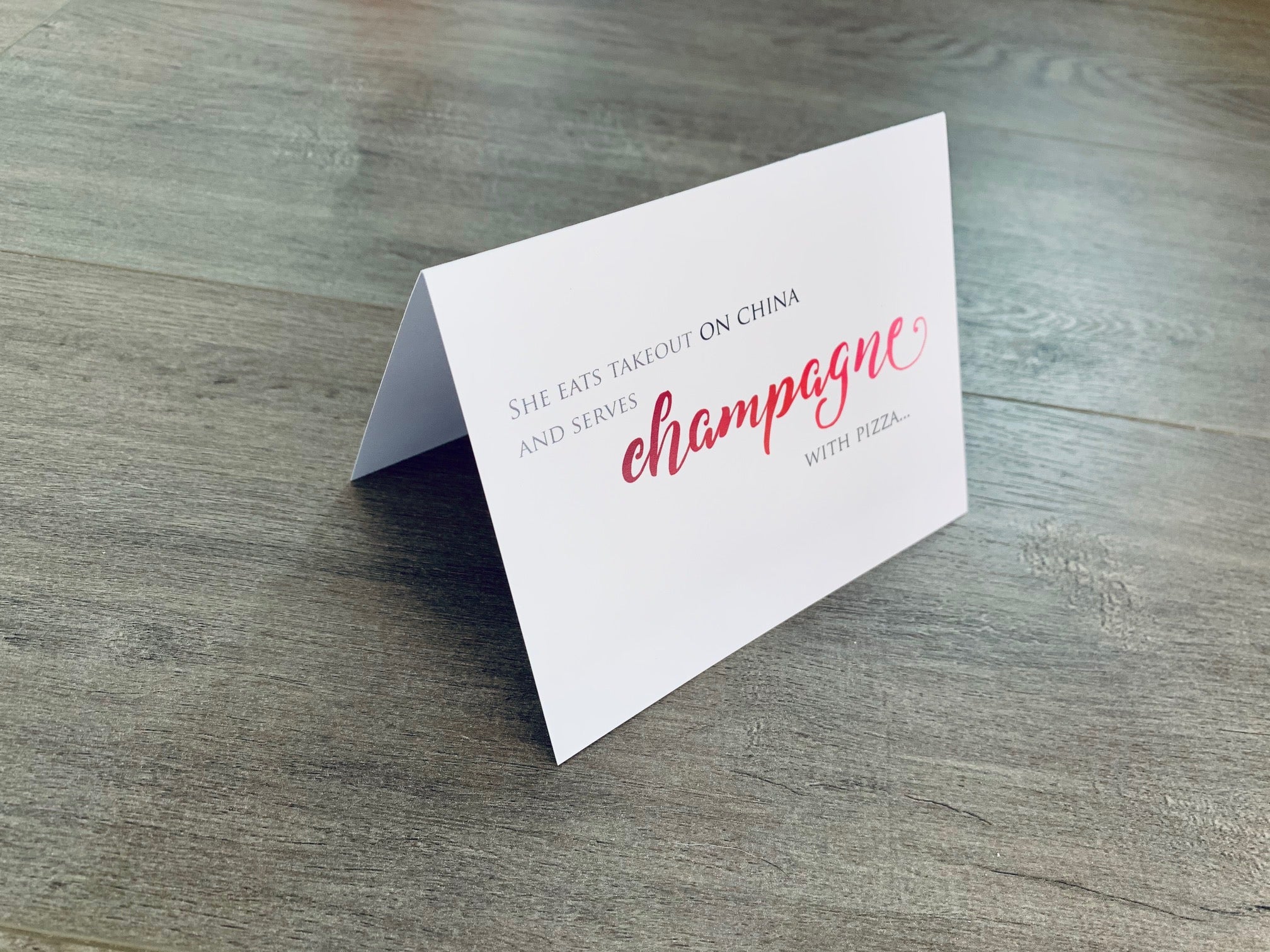 A folded white notecard is propped up on a gray wood floor. The card says, "She eats takeout on china and serves champagne with pizza." Champagne Lovers collection by Stationare.