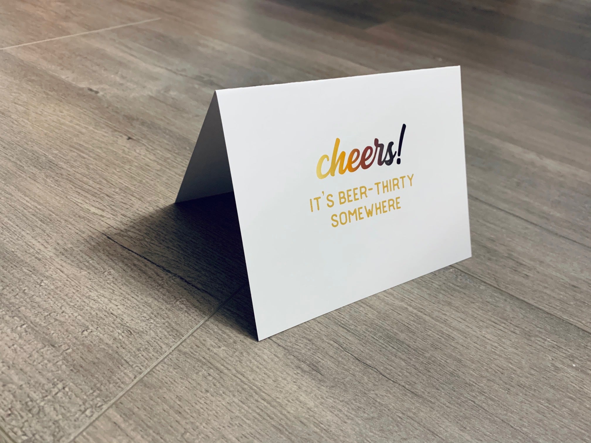 A white card is propped on a gray wood floor. The card reads, "Cheers! It's beer-thirty somewhere." Beer Lovers Collection by Stationare.
