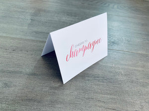 A white notecard is on a gray wooden floor. The card says, "Cheers to champagne." Champagne Lovers collection by Stationare.