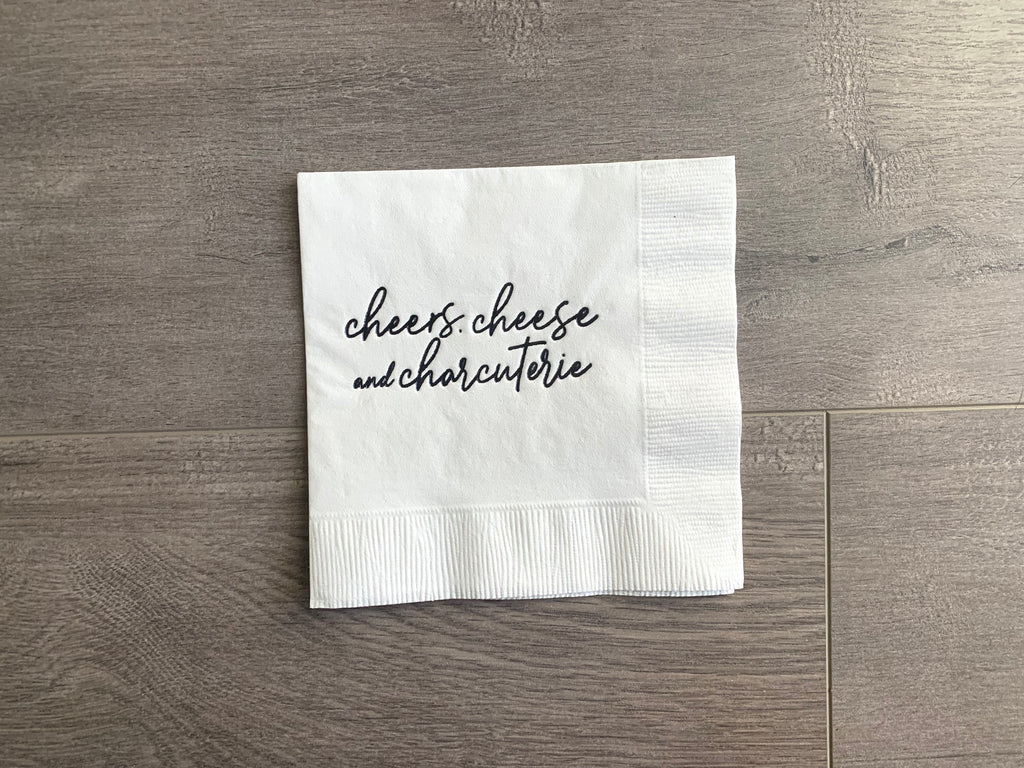 white cocktail napkin with black script printing that reads "cheers, cheese and charcuterie", sitting on a gray wooden background