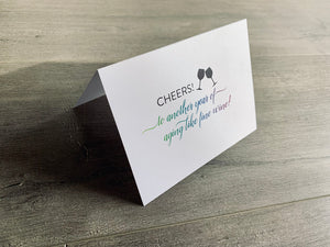A white folded notecard is propped up on a gray wooden floor. The card reads, "Cheers! to another year of aging like fine wine!" Birthday Cheers collection by Stationare