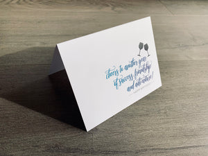 A white folded notecard is propped up on a gray wood floor. The card reads, "cheers to another year of success friendship and adventure! Happy Birthday!" Birthday card by Stationare
