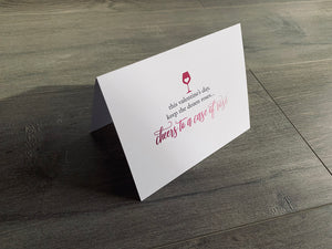A folded white notecard is propped up on a wooden floor. The card says, "This Valentine's Day, keep the dozen roses... cheers to a case of rosé" with a heart inside a wine glass. Stationare's Valen-wine collection.