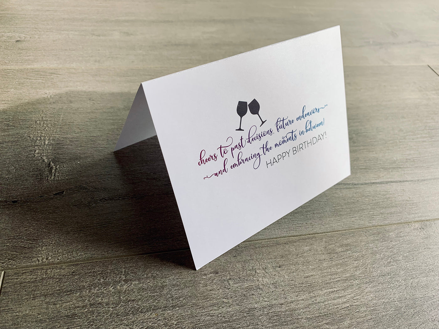 A white birthday card is folded and propped up on a wooden background. Under two dark gray wine glasses is script font that reads, "cheers to past decisions, future endeavors...and embracing the moments in-between! Happy Birthday!" Stationare's Birthday Cheers collection.