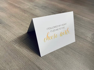 A white notecard is on a gray wooden floor. The card says, "I followed by heart. It led me to the cheese aisle." Cheese Lovers collection by Stationare.