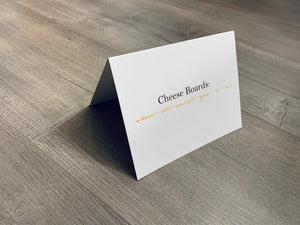 A white notecard is on a gray wooden floor. The card says, "Cheese boards - where self control goes to die." Cheese Lovers collection by Stationare.