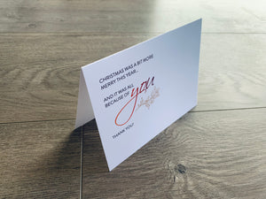A white folded notecard sits on a wooden floor. On the front of the card, it reads, "Christmas was a bit more merry this year... and it was all because of you. Thank you!" Merry Thanks Collection by Stationare.