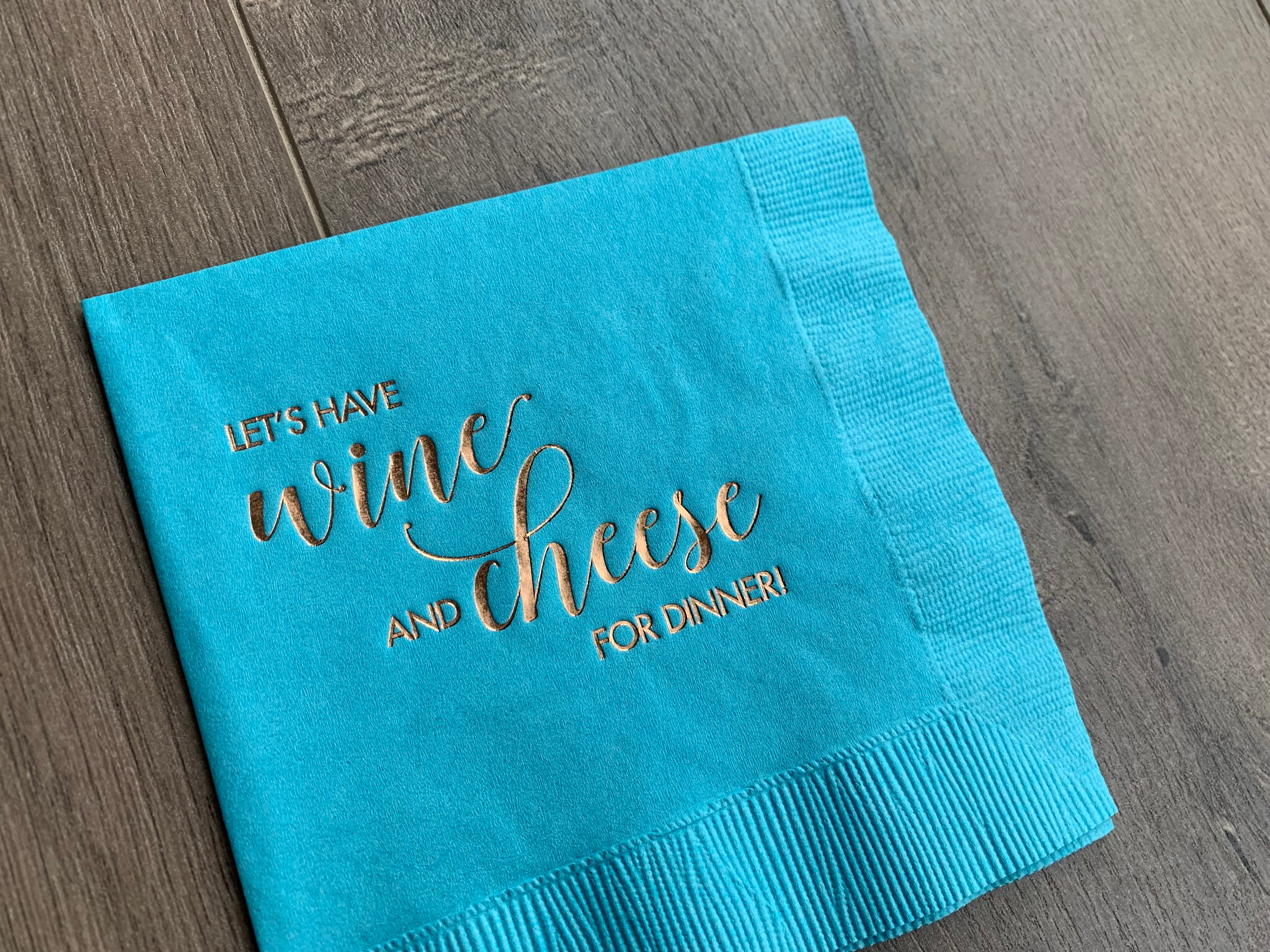 Angled closeup of a bright blue napkin with metallic gold foil that reads - Let's have wine and cheese for dinner! The napkin is slanted and slightly cut off in the frame, as it lies on a grayish wooden background.