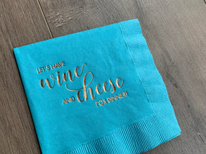 Angled closeup of a bright blue napkin with metallic gold foil that reads - Let's have wine and cheese for dinner! The napkin is slanted and slightly cut off in the frame, as it lies on a grayish wooden background.