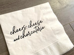 closeup of the black matte printing that reads - cheers, cheese and charcuterie - on a white cocktail napkin. It sits on a gray wooden background.