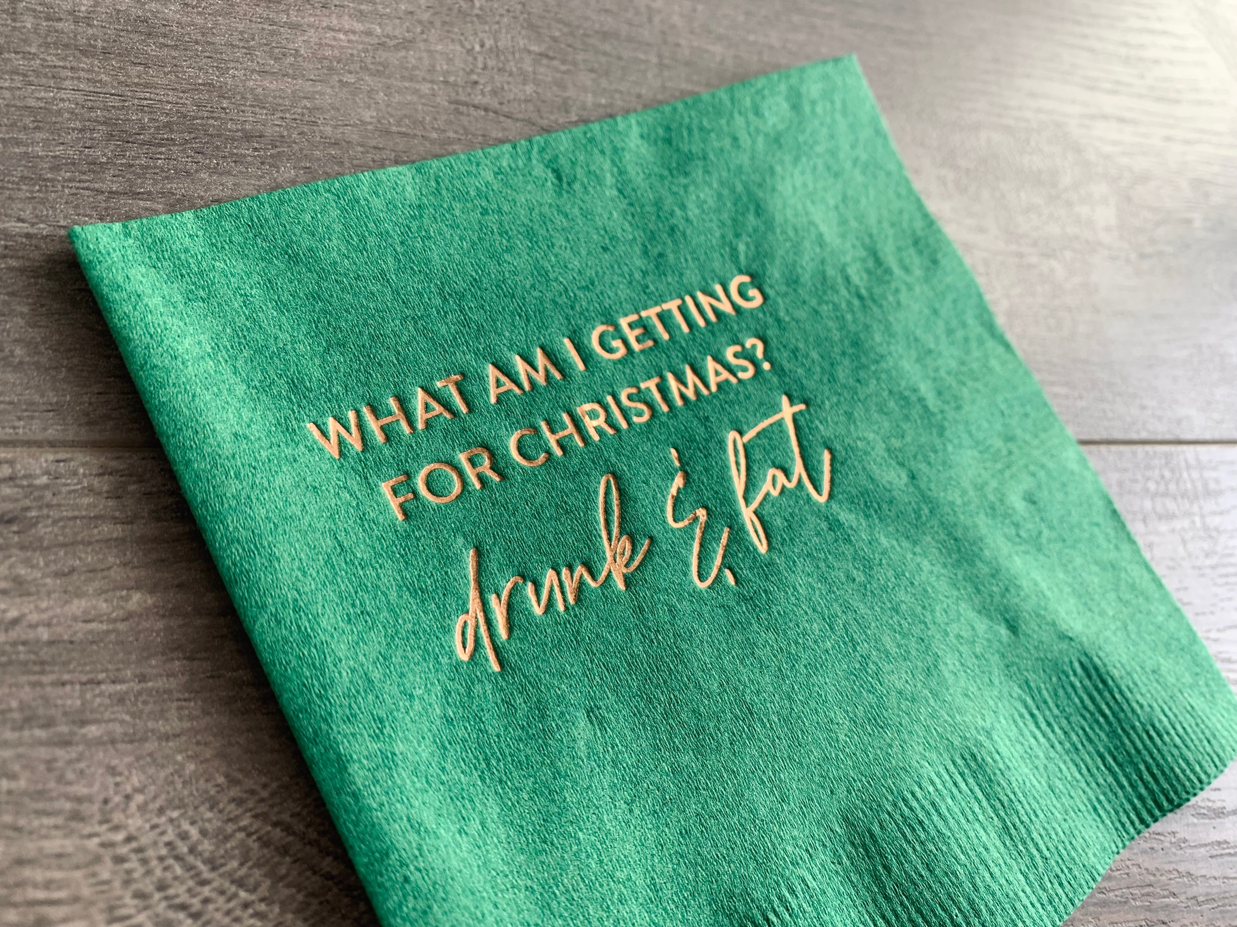 Closeup of a green cocktail napkin, angled against a gray wooden background. In gold foil, it reads: "What am I getting for Christmas? Drunk & Fat" by Stationare