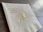 Close-up of an angled off-white cocktail napkin lies on a gray wood background. In champagne foil it reads, "Let me just tell you, the tree isn't the only thing that's going to get lit this Christmas." By Stationare