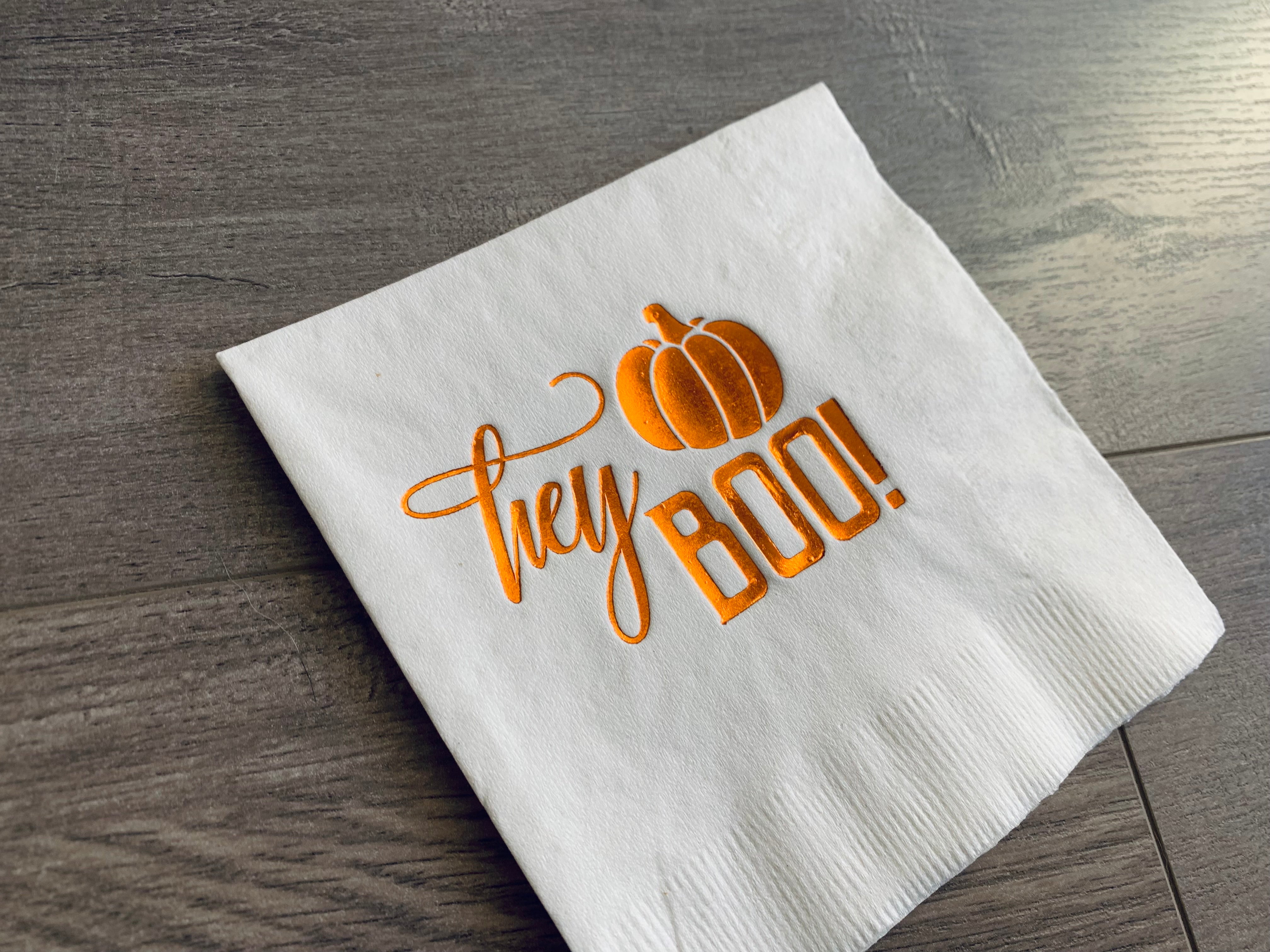 Closeup view of a white cocktail napkin with shiny, orange foil that reads, "hey Boo." A small orange pumpkin sits above the word, boo. 