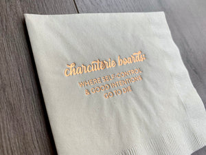 Angled image of an ivory cocktail napkin with metallic gold foil that reads - Charcuterie Boards: where self-control and good intentions go to die. The napkin is against a grayish wooden background. By Stationare.