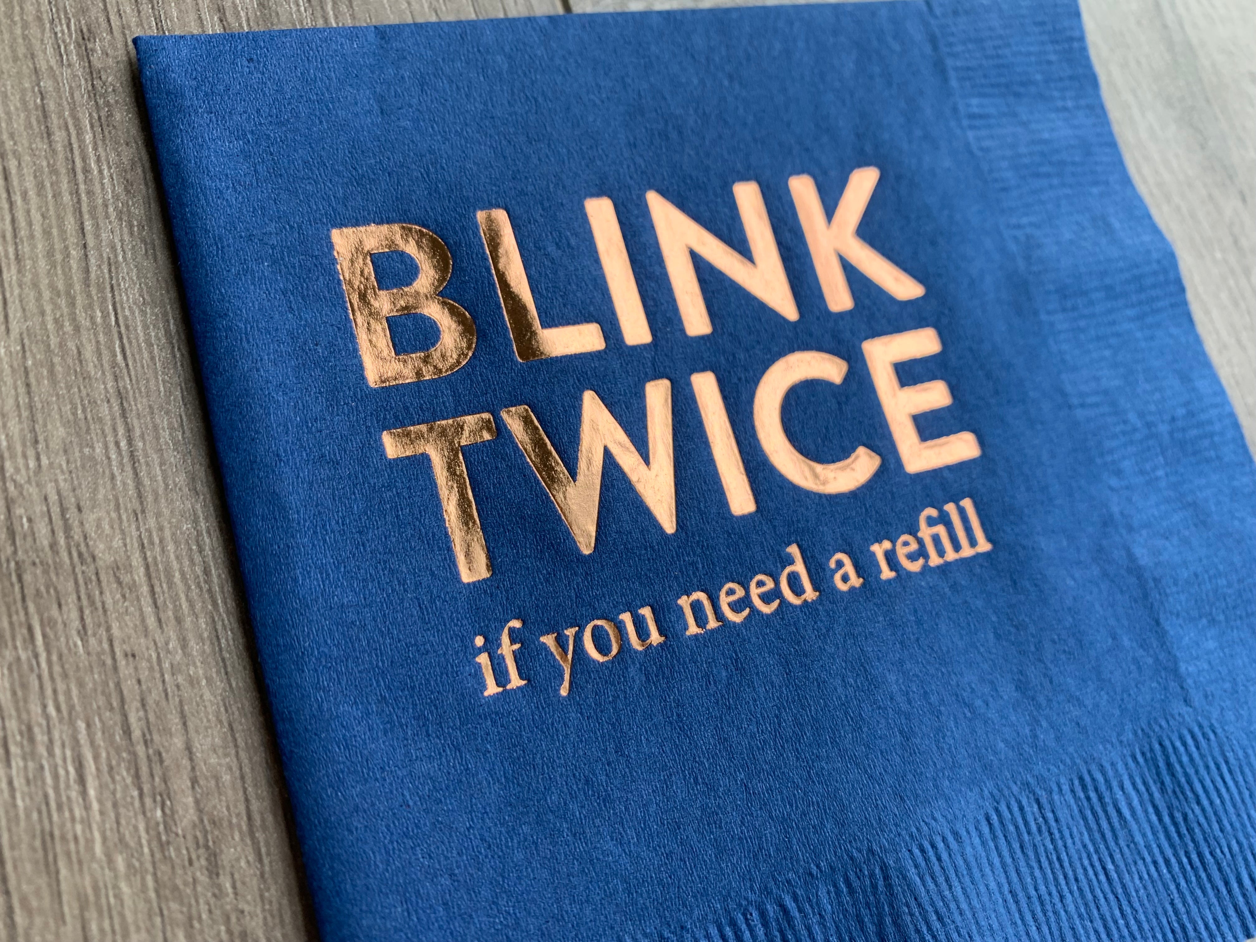 closeup of the metallic gold foil on a navy cocktail napkin that reads - blink twice if you need a refill. The napkin is slightly angled while lying on a gray wooden bakcground.