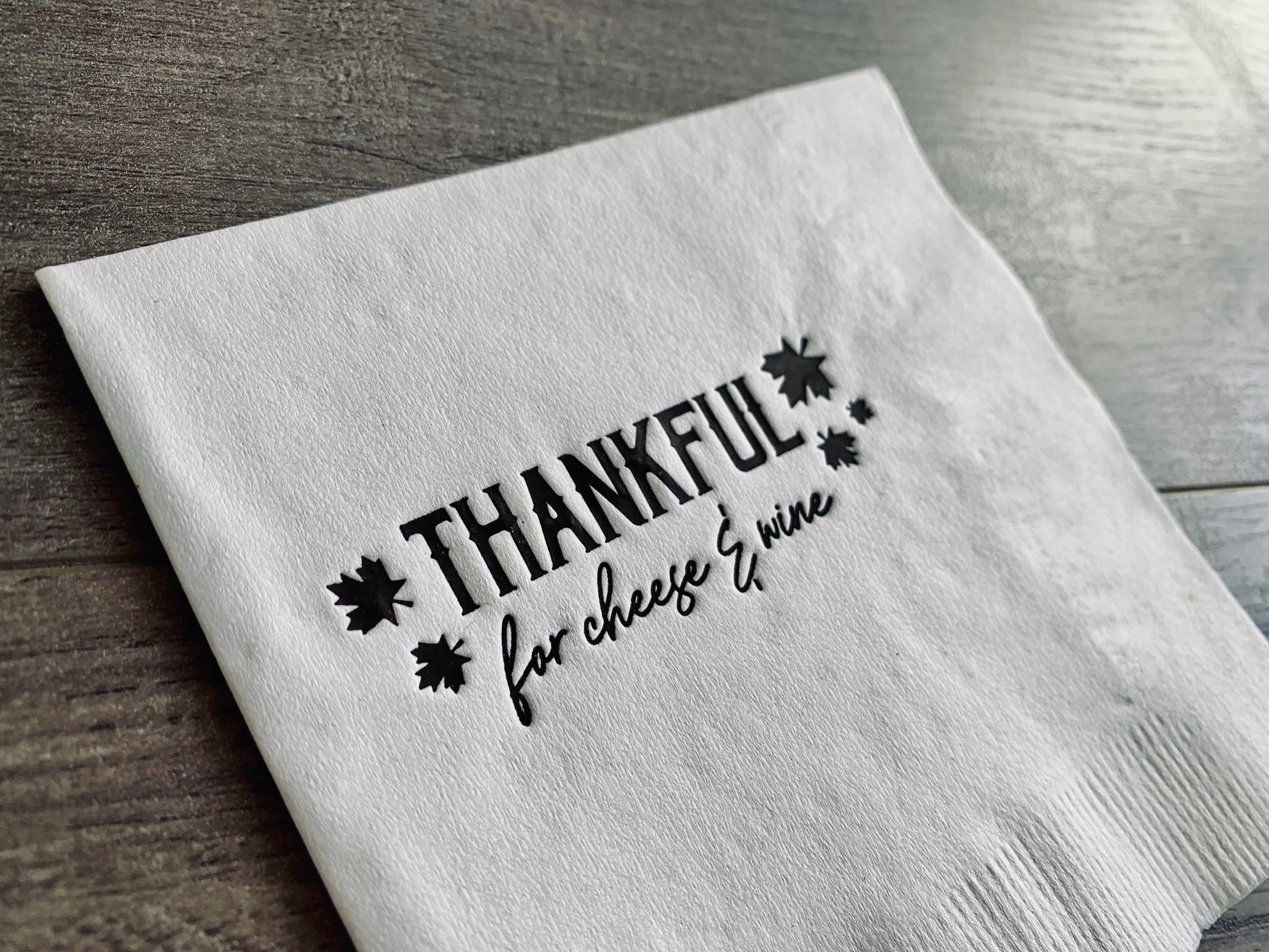Closeup of a white cocktail napkin on a gray wooden background. In brown matte foil, it reads "Thankful for cheese and wine" with small leaves. By Stationare.