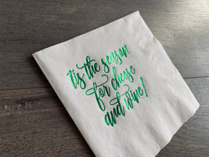 Closeup of an angled white napkin on a gray wooden background. On the napkin, in script font, it reads, "tis the season for cheese and wine." By Stationare.
