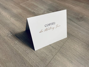 A white, folded notecard sits on a wooden floor. The card says, "Coffee = aka Adulting Juice" Coffee and Conquer collection by Stationare.