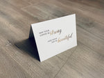 A white, folded notecard sits on a wooden floor. The card says, "may your coffee be strong and your day be beautiful." Coffee Lovers collection by Stationare.