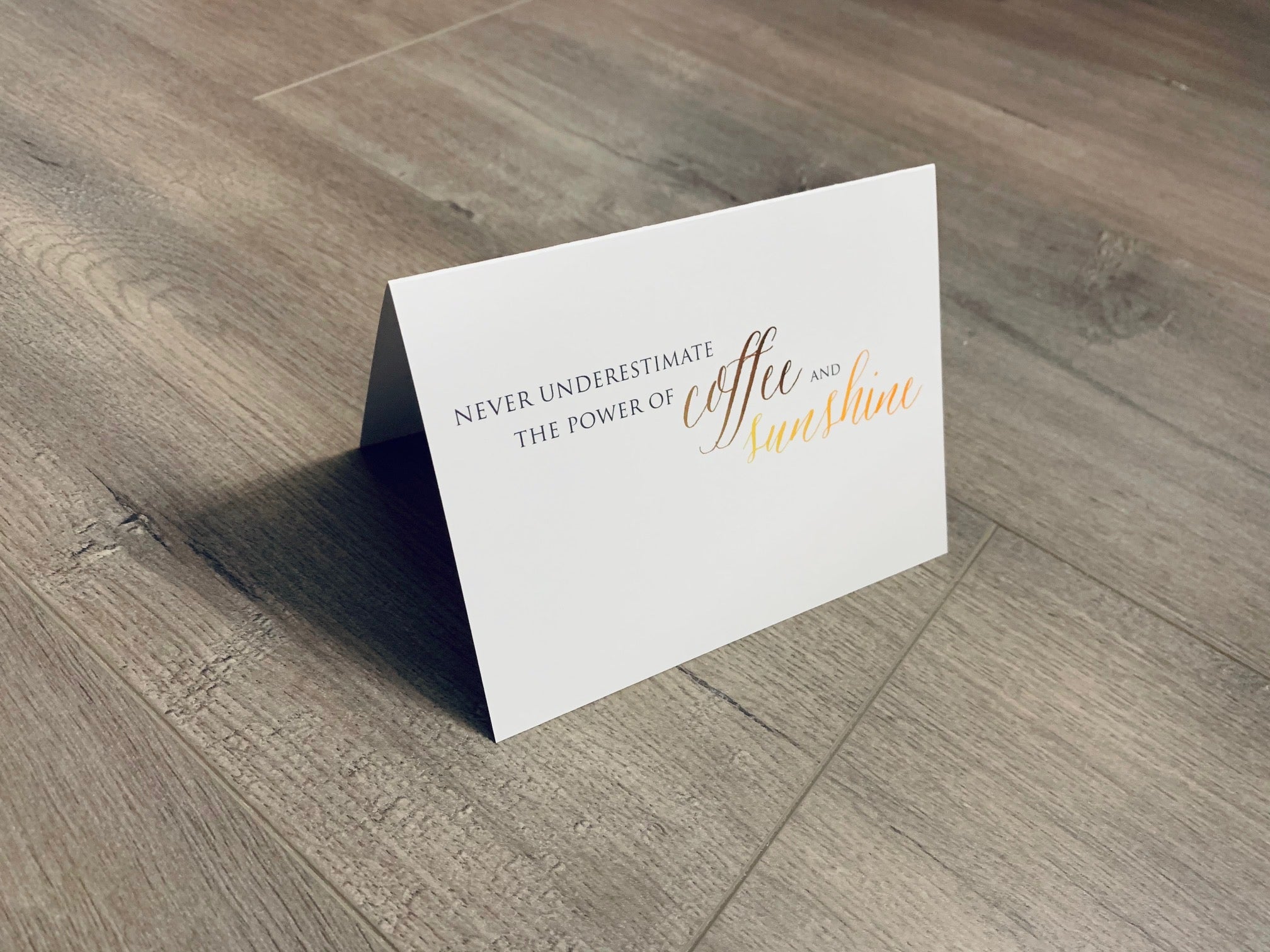 A white, folded notecard sits on a wooden floor. The card says, "Never underestimate the power of coffee and sunshine." Coffee Lovers collection by Stationare.