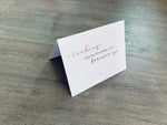 A white card is propped on a gray wood floor. The card reads, "Cookies... starting friendships since... forever ago." Bakers Collection by Stationare.