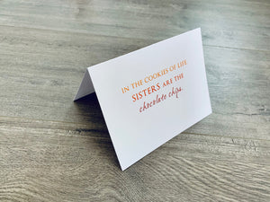 A folded white notecard is propped up on a wooden floor. The card reads, "In the cookies of life, sisters are the chocolate chips." Stationare's Sisters collection.