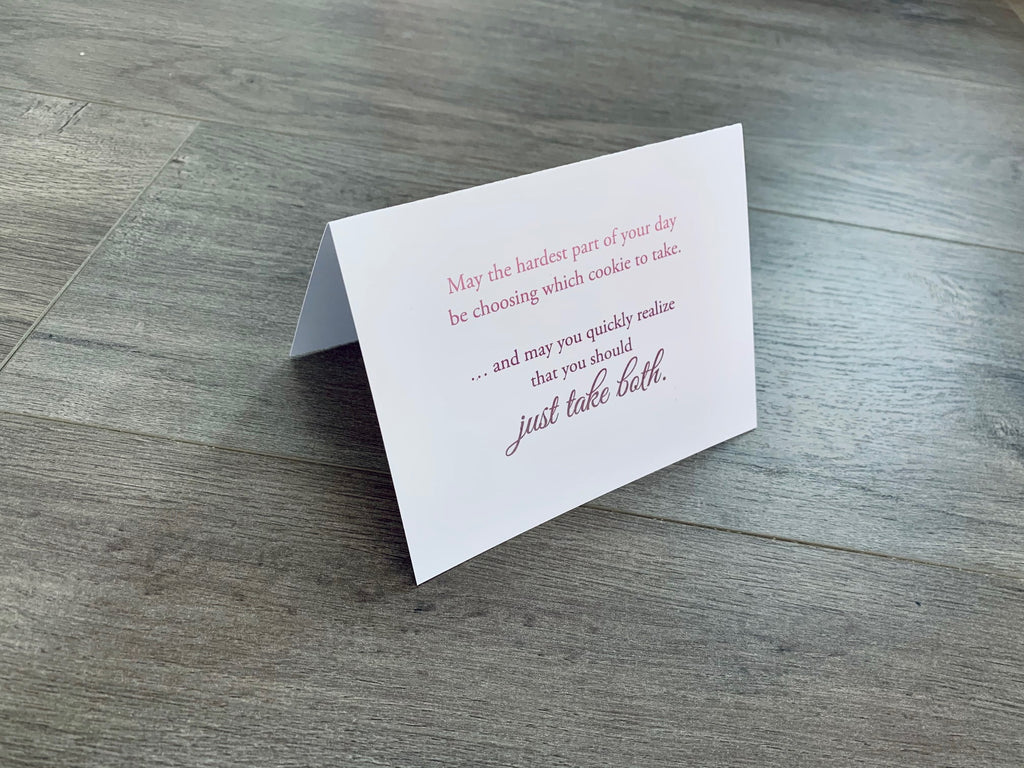 A white card is propped on a gray wood floor. The card reads, "May the hardest part of your day be choosing which cookie to take... and may you quickly realize that you should just take both." Bakers Collection by Stationare.