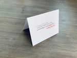 A white, folded notecard sits on a wooden floor. The card says, "Few things in the world are better than opening a bottle of wine and cooking a meal together with friends." The Foodie collection by Stationare.