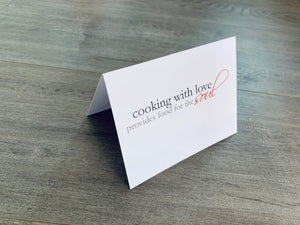 A white, folded notecard sits on a wooden floor. The card says, "Cooking with love provides food for the soul." The Foodie collection by Stationare.