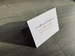 A white card is propped on a gray wood floor. The card reads, "to another year of cracking up over nothing together! Happy birthday to my partner-in-crime!" birthday card by Stationare