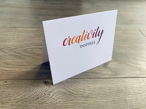 A white, folded notecard sits on a wooden floor. The card says, "Creativity inspires." Creativity collection by Stationare.