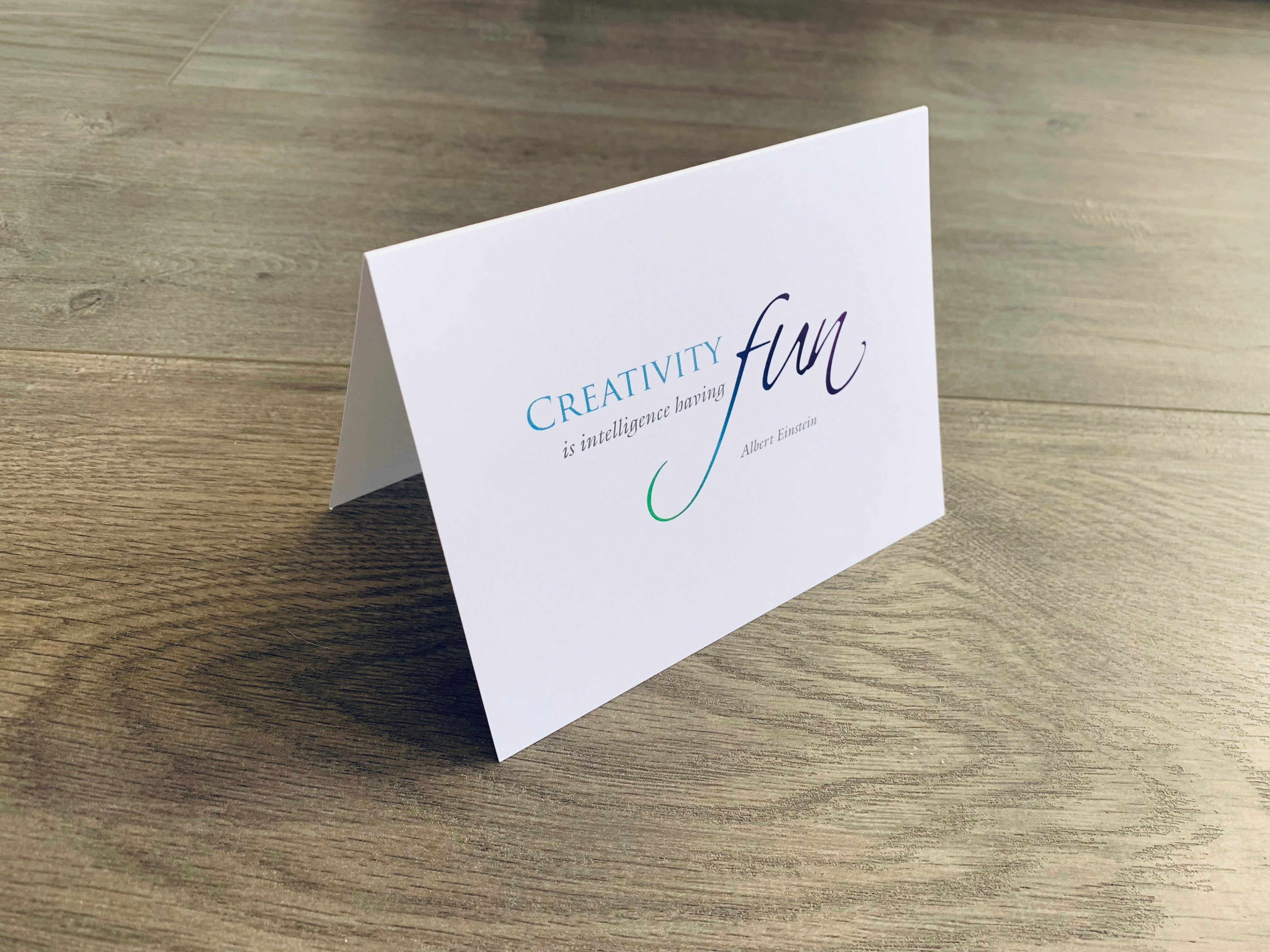 A white, folded notecard sits on a wooden floor. The card hosts an Albert Einstein quote. It says, "Creativity is intelligence having fun." Creativity collection by Stationare.