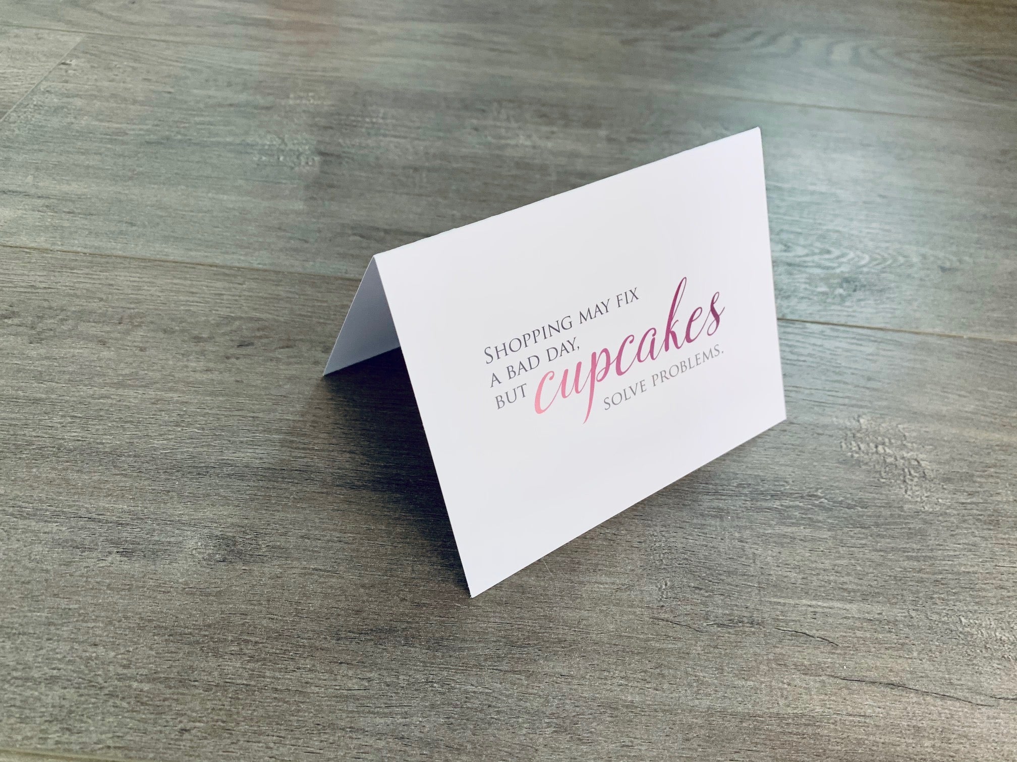 A white card is propped on a gray wood floor. The card reads, "Shopping may fix a bad day, but cupcakes solve problems." Bakers Collection by Stationare.
