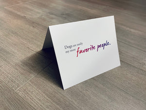 A white, folded notecard sits on a wooden floor. The card says, "Dogs are easily my most favorite people." Dog Lovers collection by Stationare.