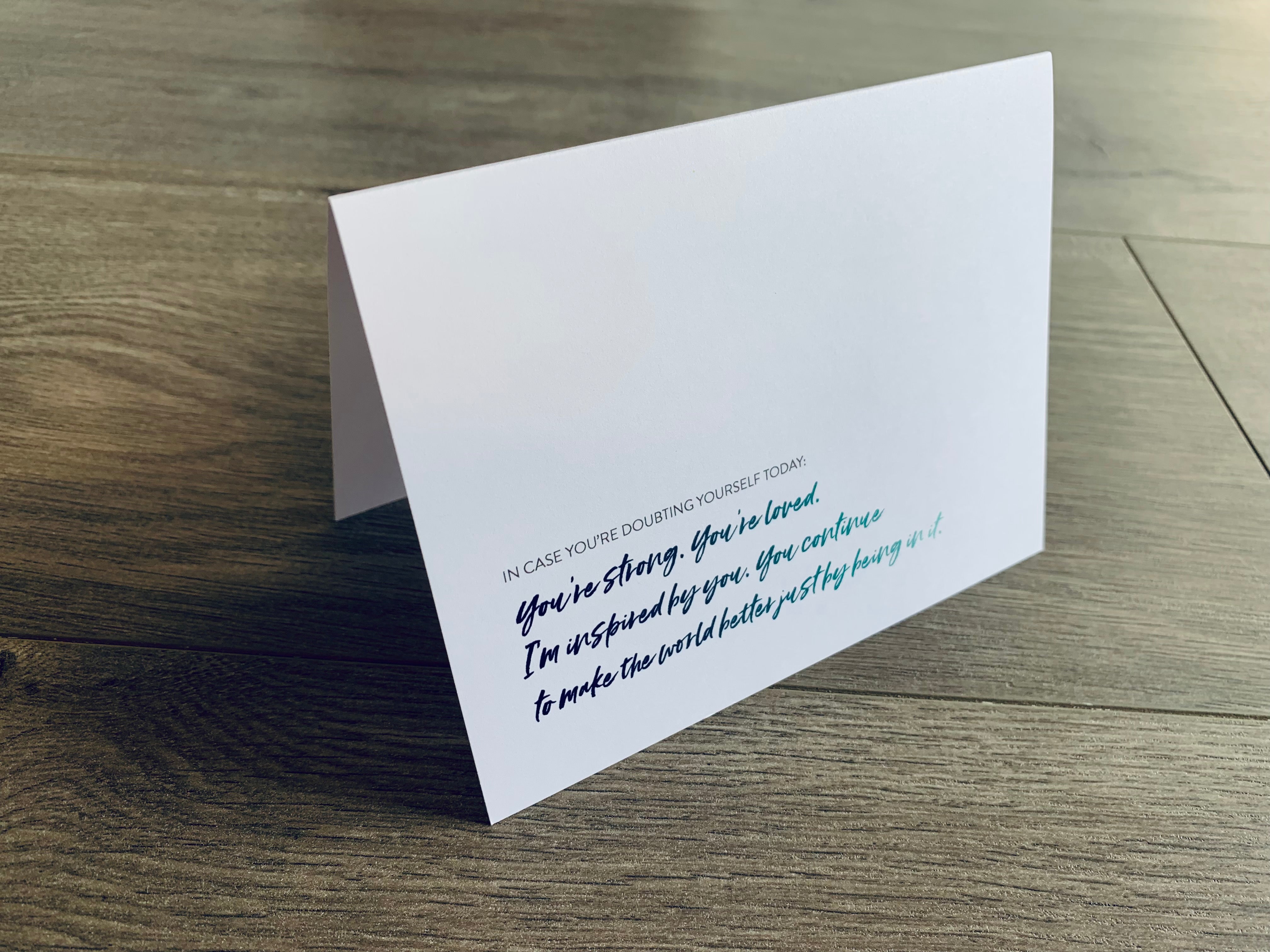 A white, folded notecard is propped up on a gray wooden floor. On the front of the card, it reads, "In case you're doubting yourself today: You're strong. You're loved. I'm inspired by you. You continue to make the world better just by being in it." 