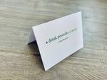 A white folded notecard is propped up on a wooden floor. The card has the Irish proverb, "a drink precedes a story." From the Irish Laughs Collection by Stationare.