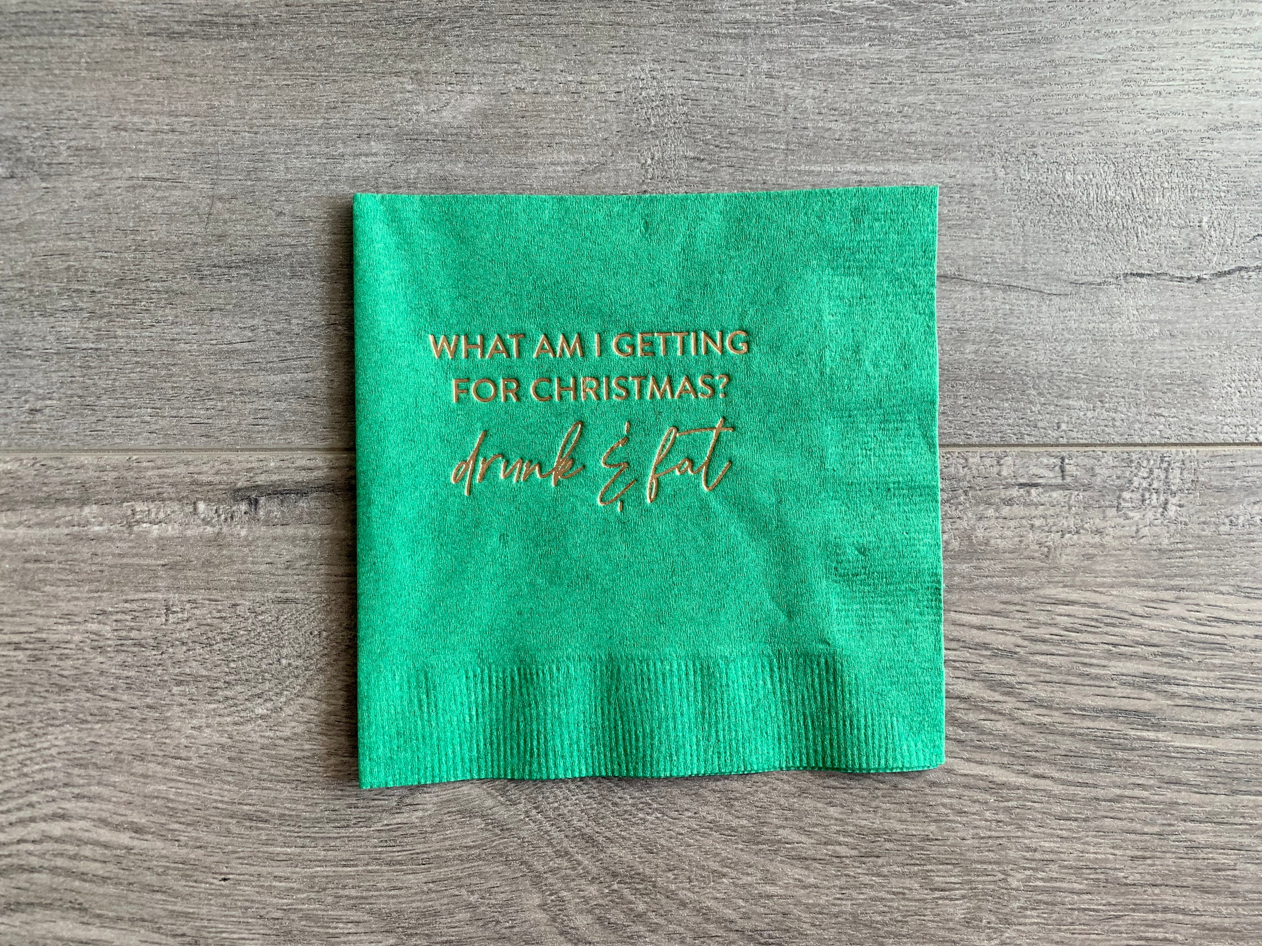 Green Christmas-themed cocktail napkin sits on a gray wooden background. In champagne foil it reads, "What am I getting for Christmas? Drunk & Fat." By Stationare