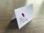 A white folded notecard stands on a gray wood floor. The card reads, "Eat. Drink. and be merry." in red and black text, along with a red champagne flute with a snowflake in it. Christmas Cheers by Stationare.