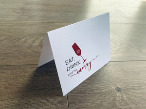 A white folded notecard stands on a gray wood floor. The card reads, "Eat. Drink. and be merry." in red and black text, along with a red champagne flute with a snowflake in it. Christmas Cheers by Stationare.