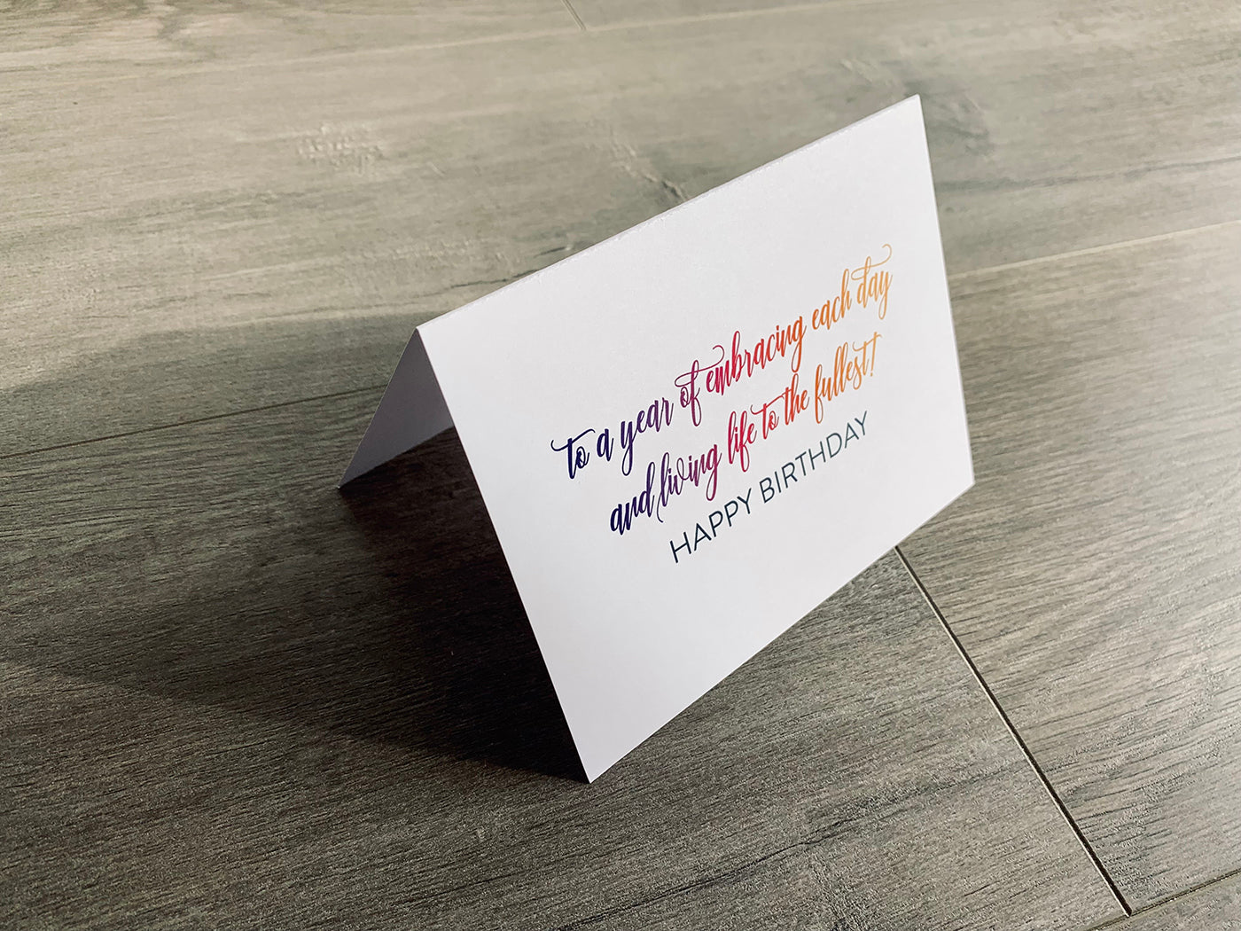 A white card is propped on a gray wood floor. The card reads, "to a year of embracing each day and living life to the fullest! Happy Birthday!"  birthday card by Stationare
