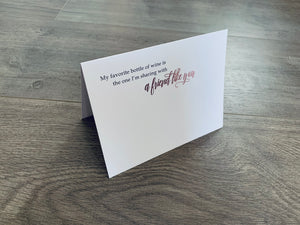 A white notecard is folded and propped up on a wooden floor. The card says, "My favorite bottle of wine is the one I'm sharing with a friend like you." Wine-themed notecards by Stationare.