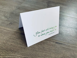 A white folded notecard sits on a gray wood floor. It reads, "Your feet will bring you to where your heart is." Irish Inspirations collection by Stationare.