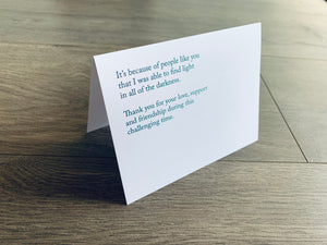 A folded white notecard is propped up on a wooden floor. The card reads, "It's because of people like you that I was able to find light in all the darkness. Thank you for your love, support and friendship during this challenging time." Stationare's Surviving Hard Times collection.