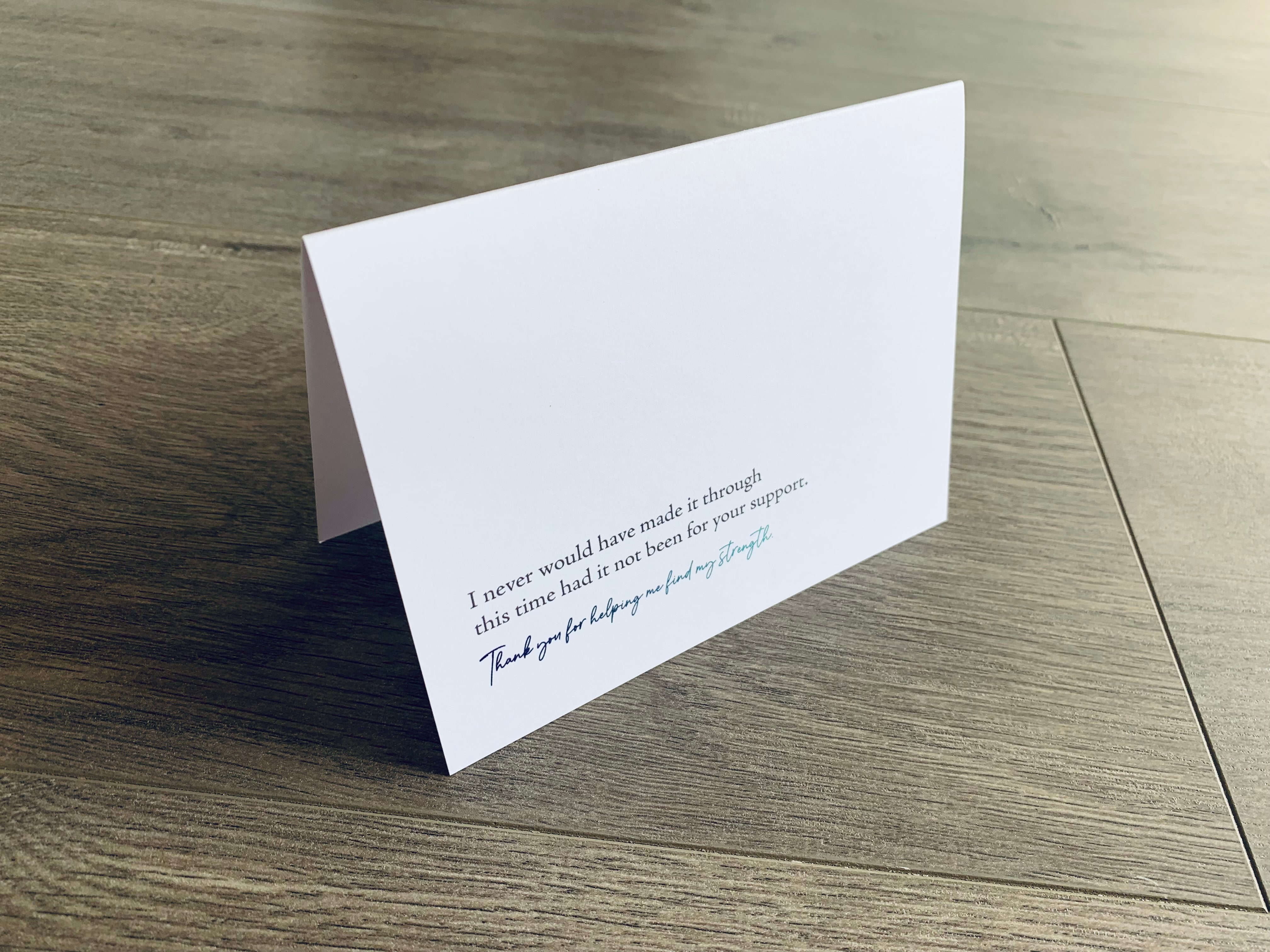 A folded white notecard is propped up on a wooden floor. The card reads, "I never would have made it through this time had it not been for your support. Thank you for helping me find my strength." Stationare's Surviving Hard Times collection.