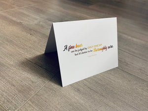 A white card is propped on a gray wood floor. The card reads, "A fine beer can be judged by only one sip but it's better to be thoroughly sure." Beer Lovers collection by Stationare.