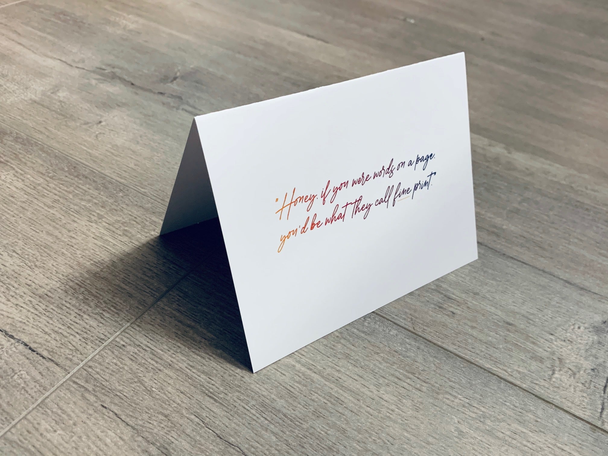 A folded white notecard is propped up on a wooden floor. The card reads, "Honey, if you were words on a page, you'd be what they call fine print." Stationare's Pick Up Lines collection.