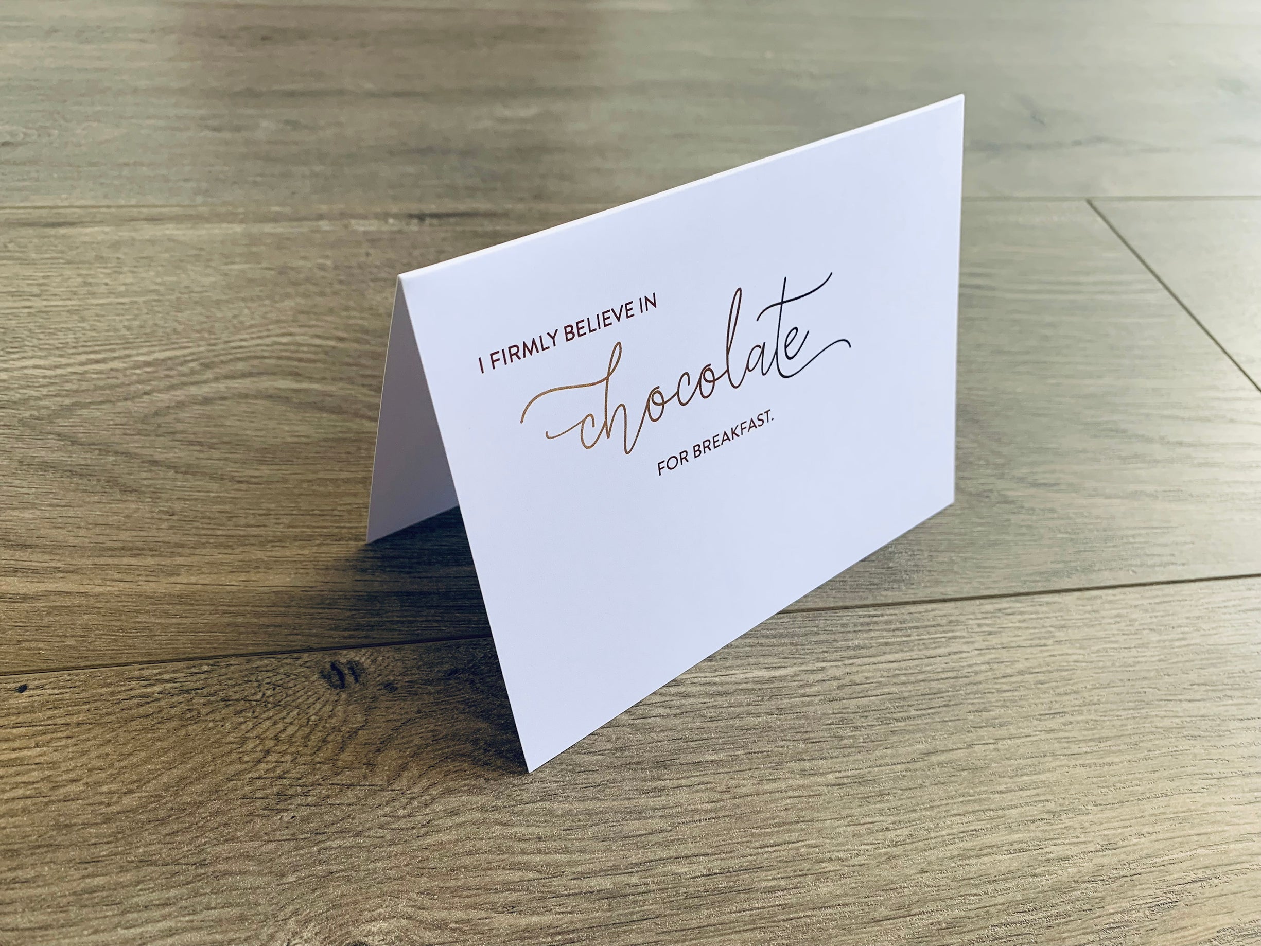 A white, folded notecard is propped up on a wooden background. The card reads "I firmly believe in chocolate for breakfast." Card by Stationare.