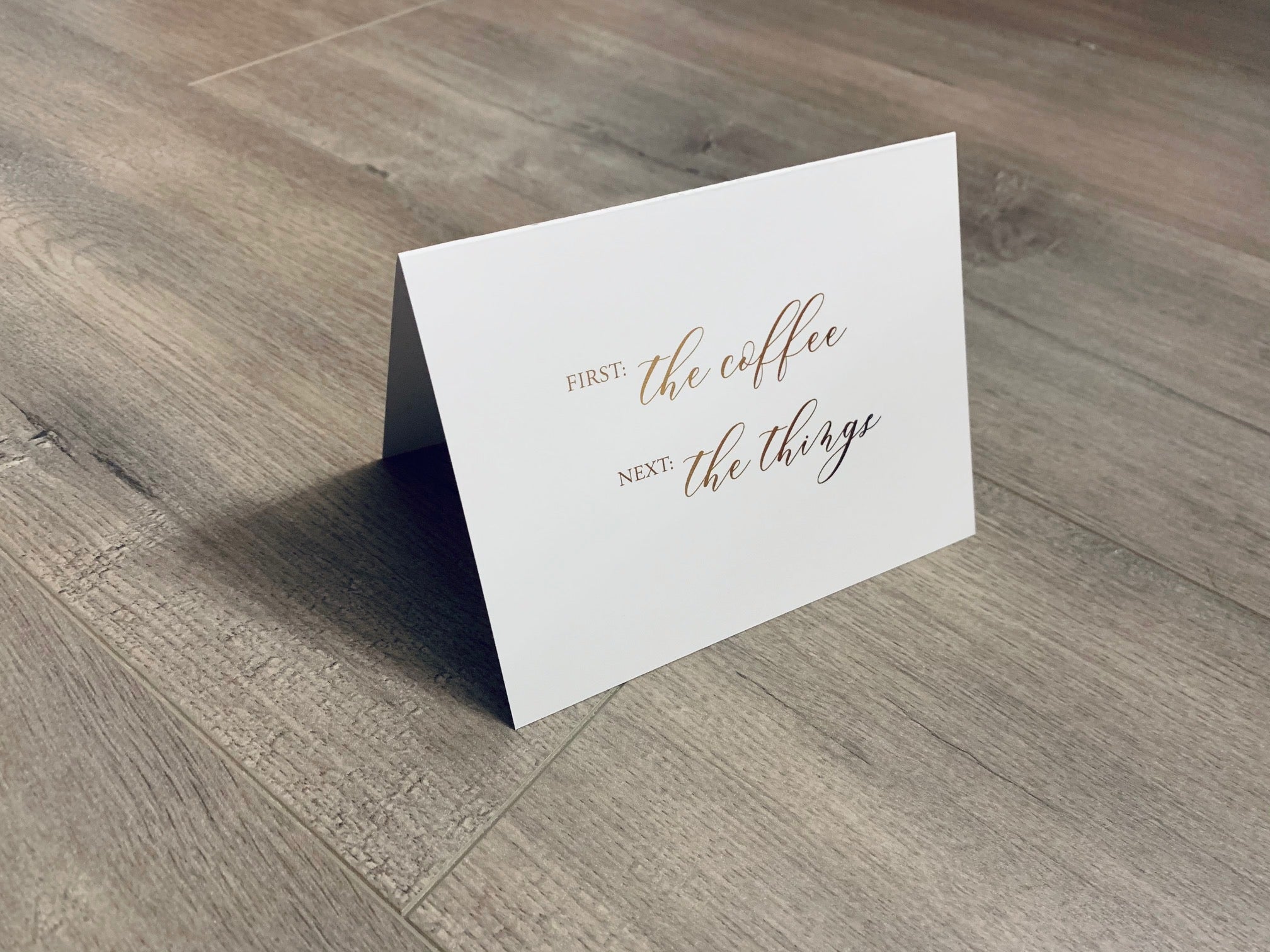 A white, folded notecard sits on a wooden floor. The card says, "First - the coffee. Next - the things." Coffee and Conquer collection by Stationare.