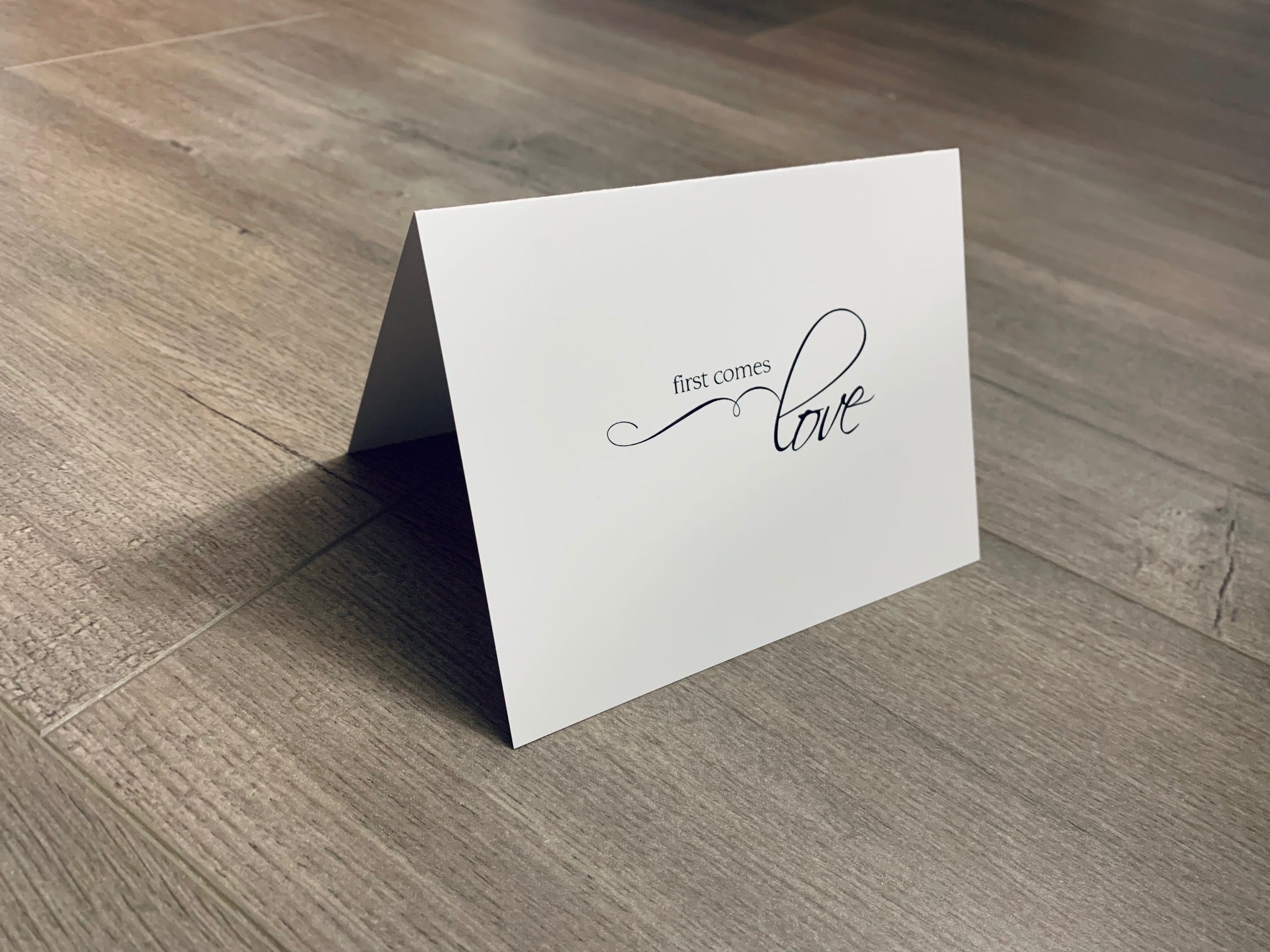 A white folded notecard is propped up on a wooden floor. The card says, "first comes love" in black ink. Stationare's Bridal and Wedding Collection.