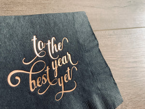 Closeup image of gold foil script printing that reads, "to the best year yet." It's printed on a black cocktail napkin with a wooden background.